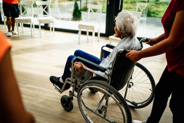 An Elderly women in a wheelchair being pushed by a caregiver in a convalescent Facility